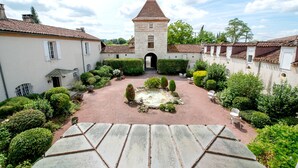Un patio con una fuente central, rodeado de setos bien cuidados y un camino de piedra que conduce a un edificio con una torre.