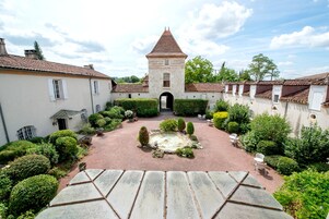 Un patio con una fuente central, rodeado de setos bien cuidados y un camino de piedra que conduce a un edificio con una torre.