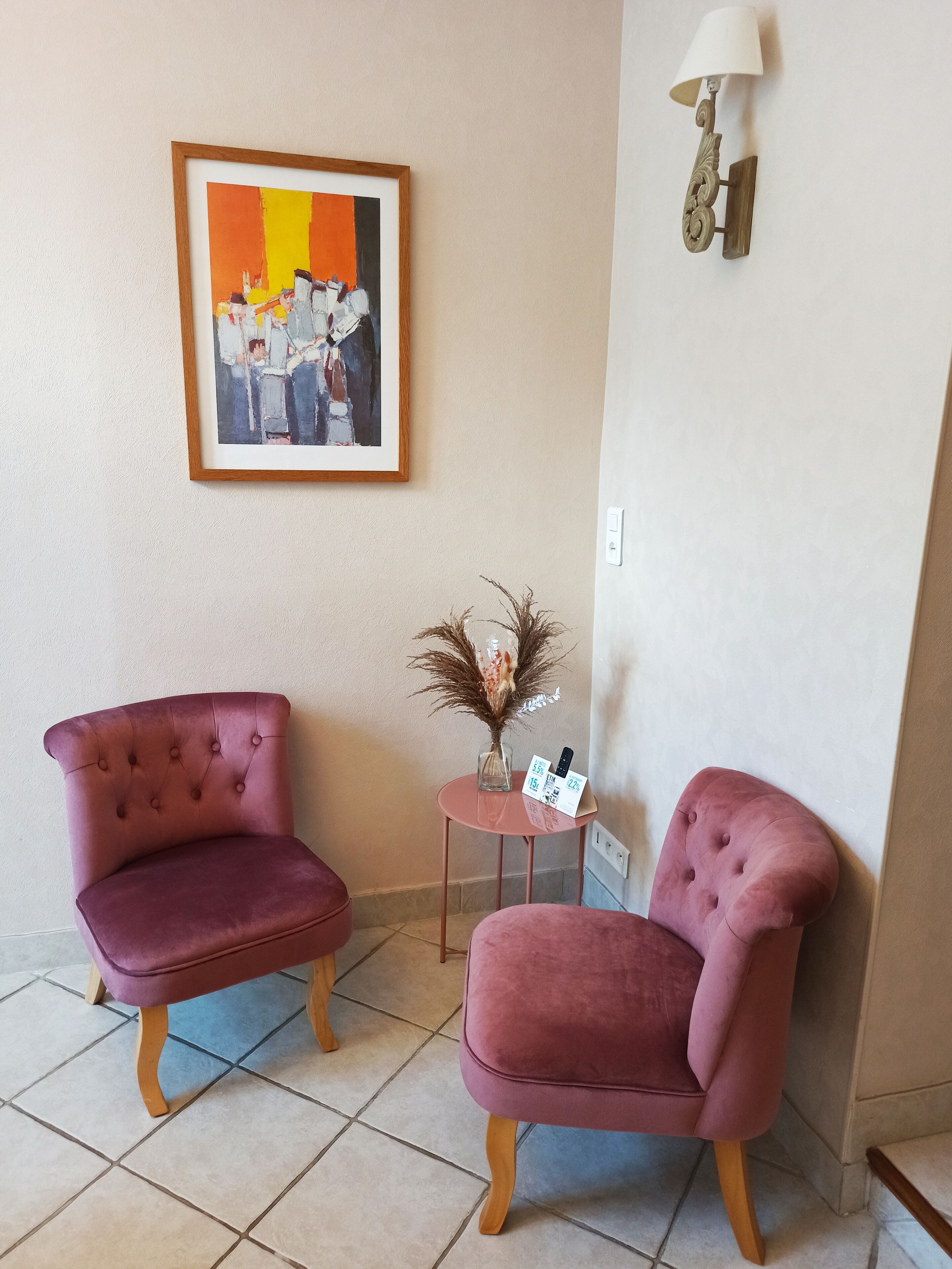 Een kamer met twee comfortabele fauteuils, een kleine tafel met een vaas en een afstandsbediening, en een lijst met een schilderij aan de muur.