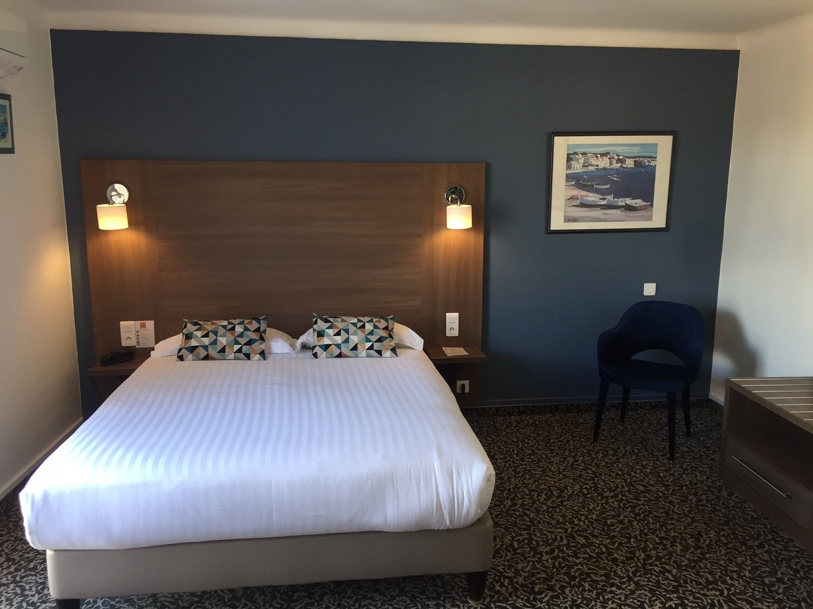 Un cuarto de hotel con una cama grande, dos lámparas de noche, una silla y un cuadro enmarcado en la pared.
