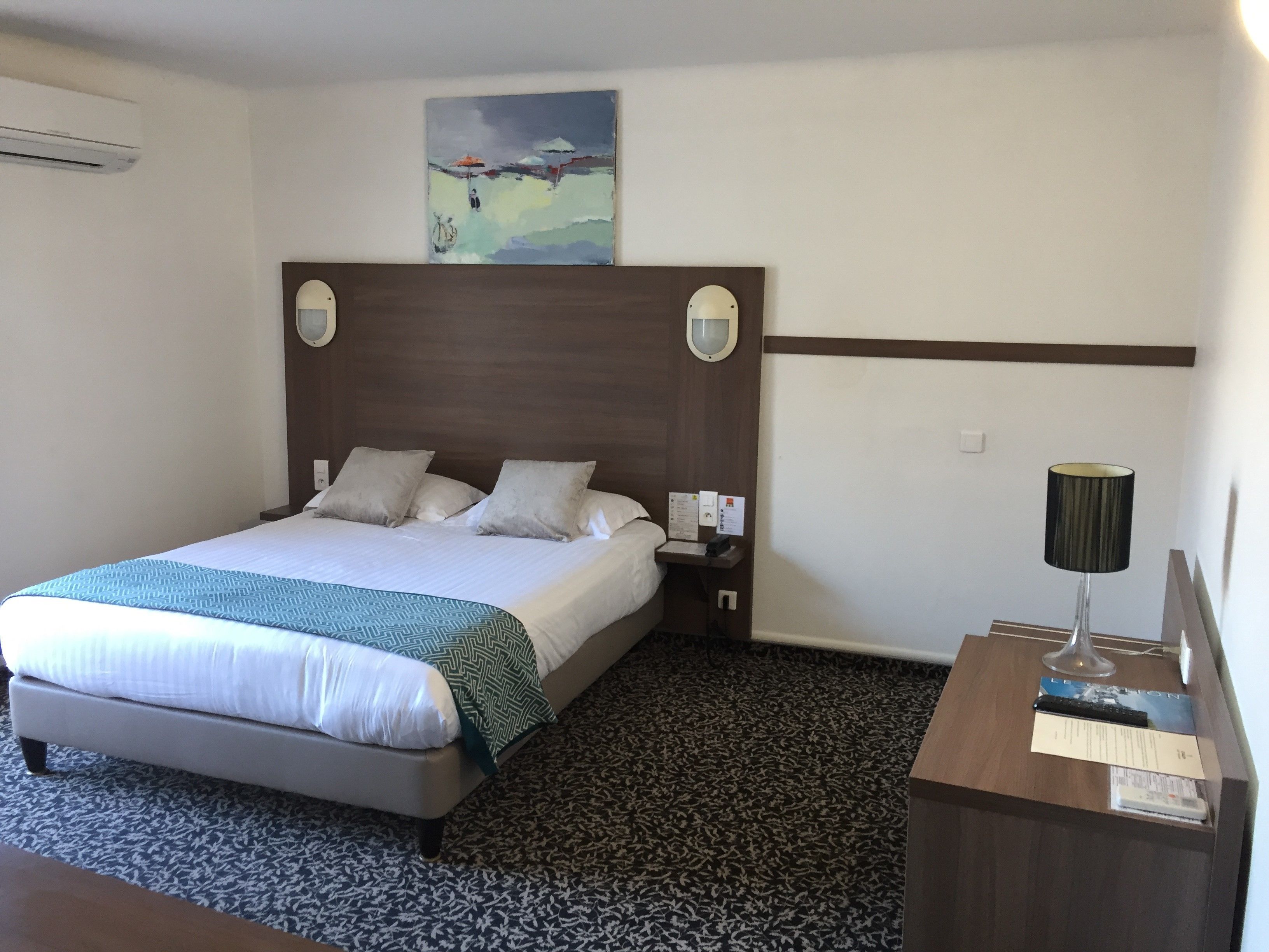 A hotel room with a large bed, a nightstand, a desk with a lamp, and a painting on the wall.