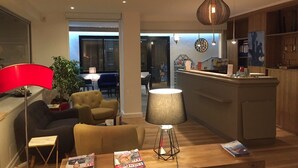 A modern living room with a sofa, a coffee table with magazines, a lamp, and a dining area with chairs and a table. - Logis Hotel et Restaurant Le Galion Canet Plage (Canet-en-Roussillon)