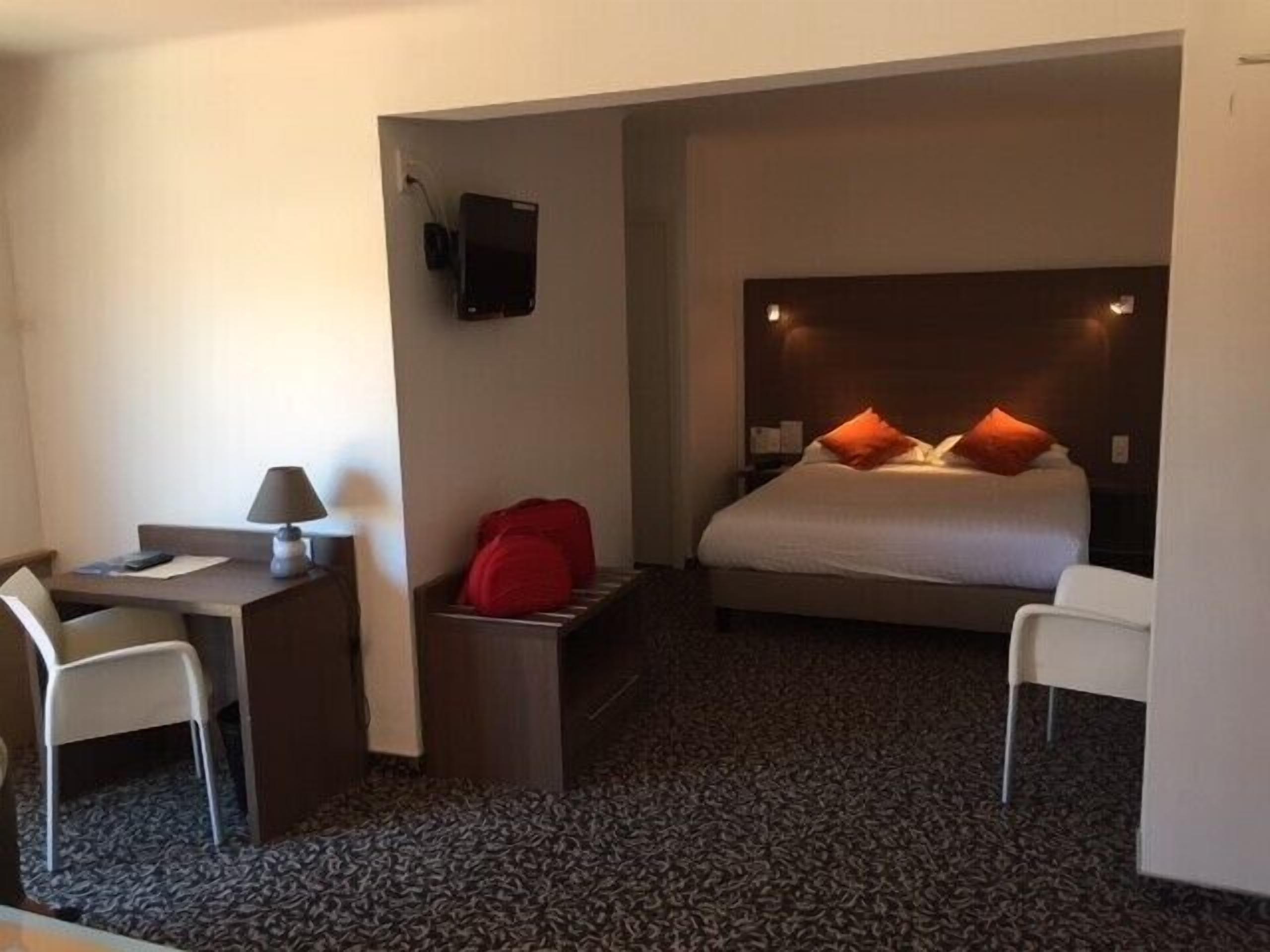 Una habitación de hotel con cama, escritorio, silla, televisor montado en la pared y una bolsa roja en el suelo.