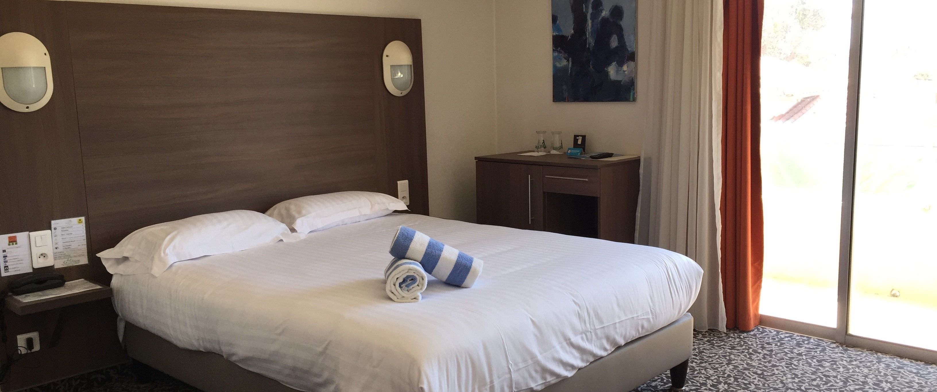 Una habitación de hotel con una cama grande, una mesita de noche con lámpara, una pequeña mesa con lámpara y un ventanal con cortinas.