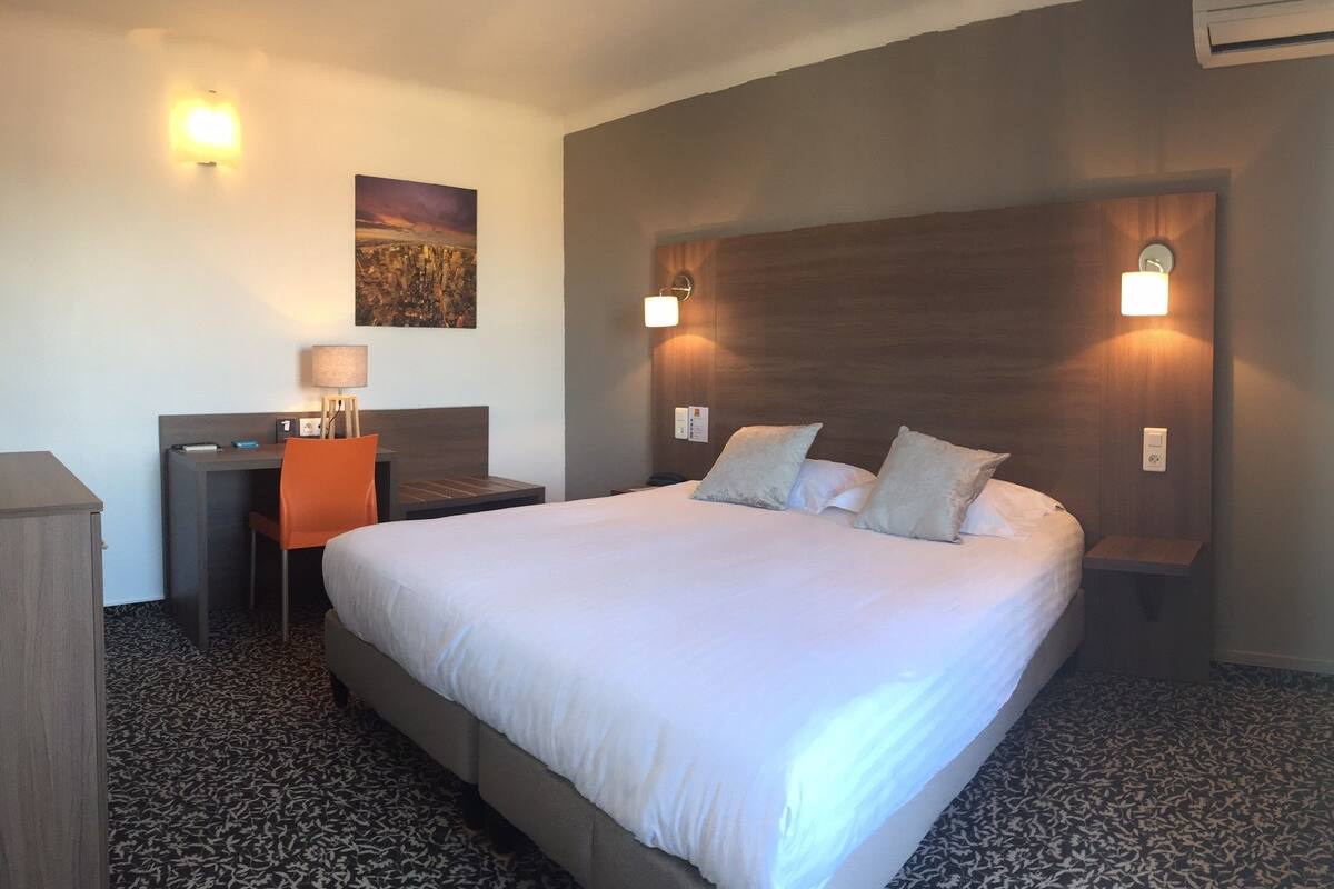 A hotel room with a large bed, a desk with a chair, a nightstand, and a wall-mounted lamp.