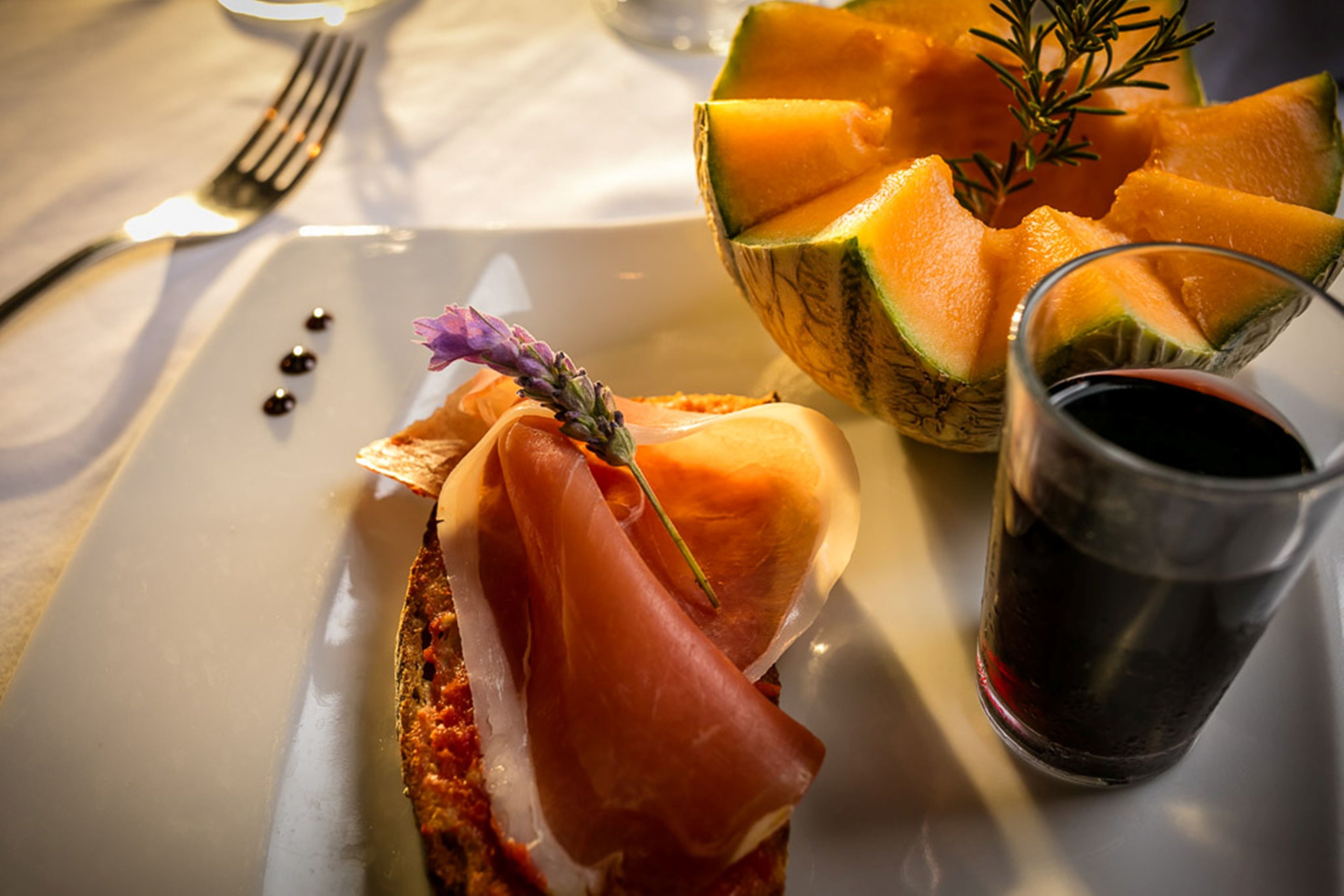 A plate with a sandwich, a glass of red wine, and a bowl of cantaloupe.