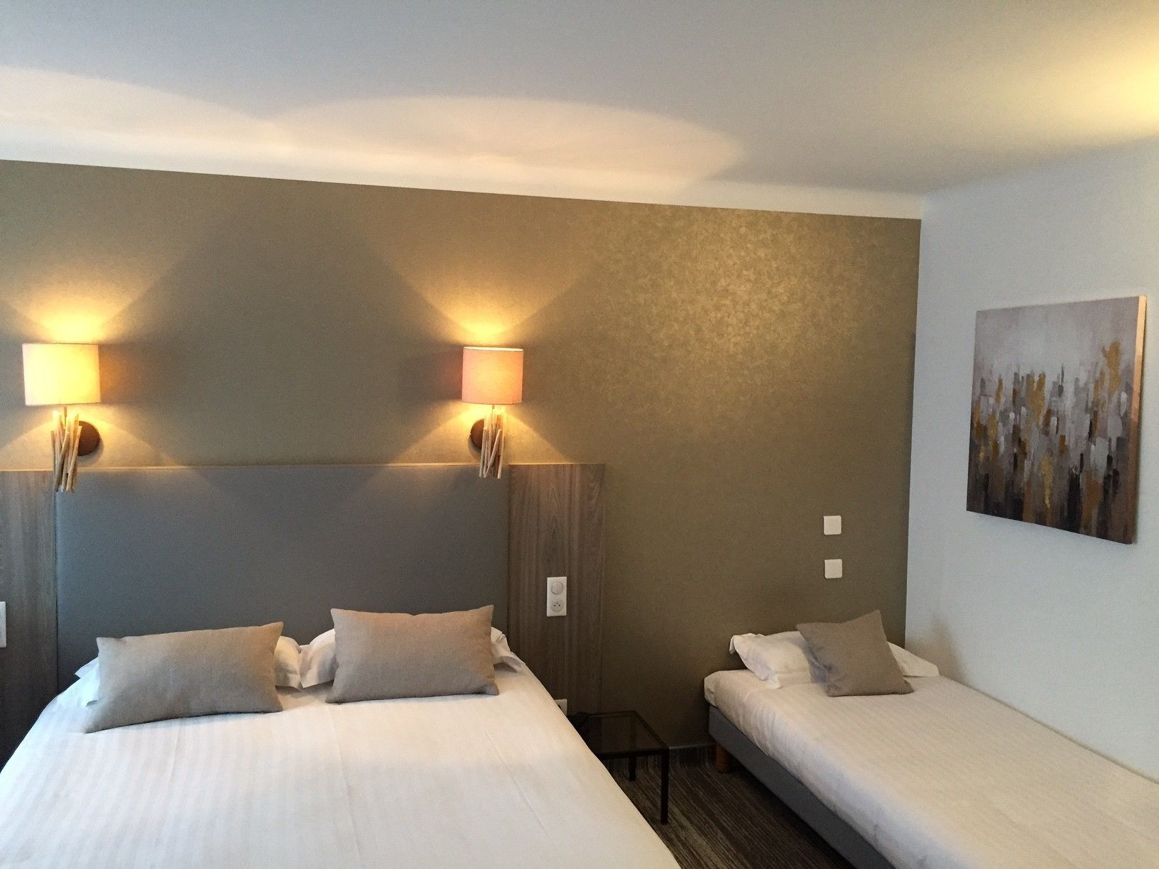 A hotel room with two beds, each with a pillow and a nightstand. There are wall-mounted lamps on either side of the beds.