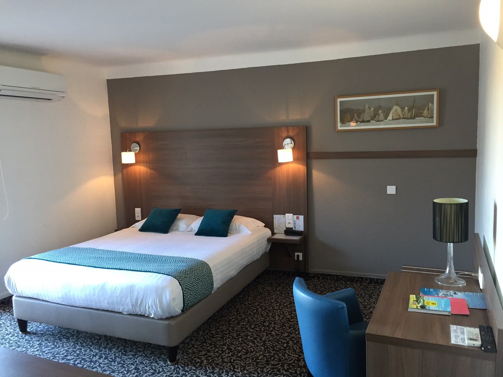 A hotel room with a bed, two bedside lamps, a desk with a lamp, a chair, and a painting on the wall.