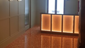 Un pasillo con suelo de mármol pulido, una recepción con paneles iluminados, un ventanal grande y un candelabro en el techo.