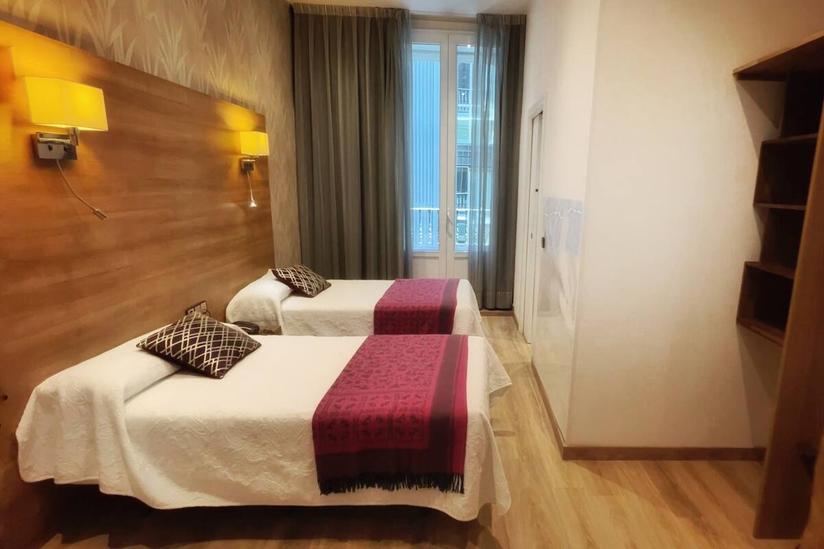 Quarto de hotel com duas camas, cabeceira de madeira, uma pequena prateleira de madeira, uma janela com cortinas e uma porta.