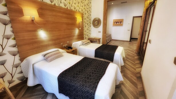 Habitación de hotel con dos camas, cabecero de madera, lámparas fijadas a la pared y un papel tapiz estampado.