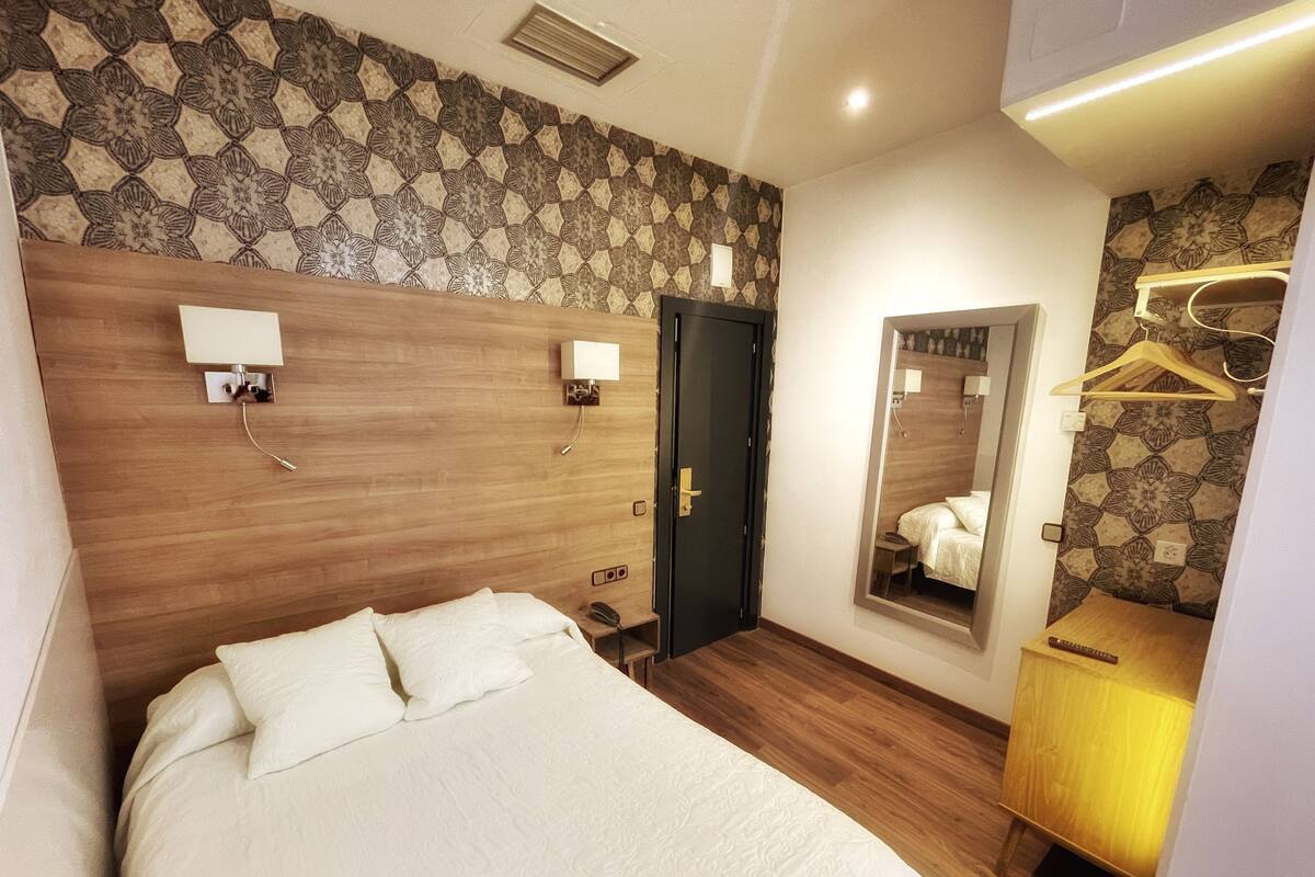 Quarto de hotel com uma cama grande, cabeceira de madeira, duas luminárias de apoio, um espelho, cômoda de madeira e um cabideiro.