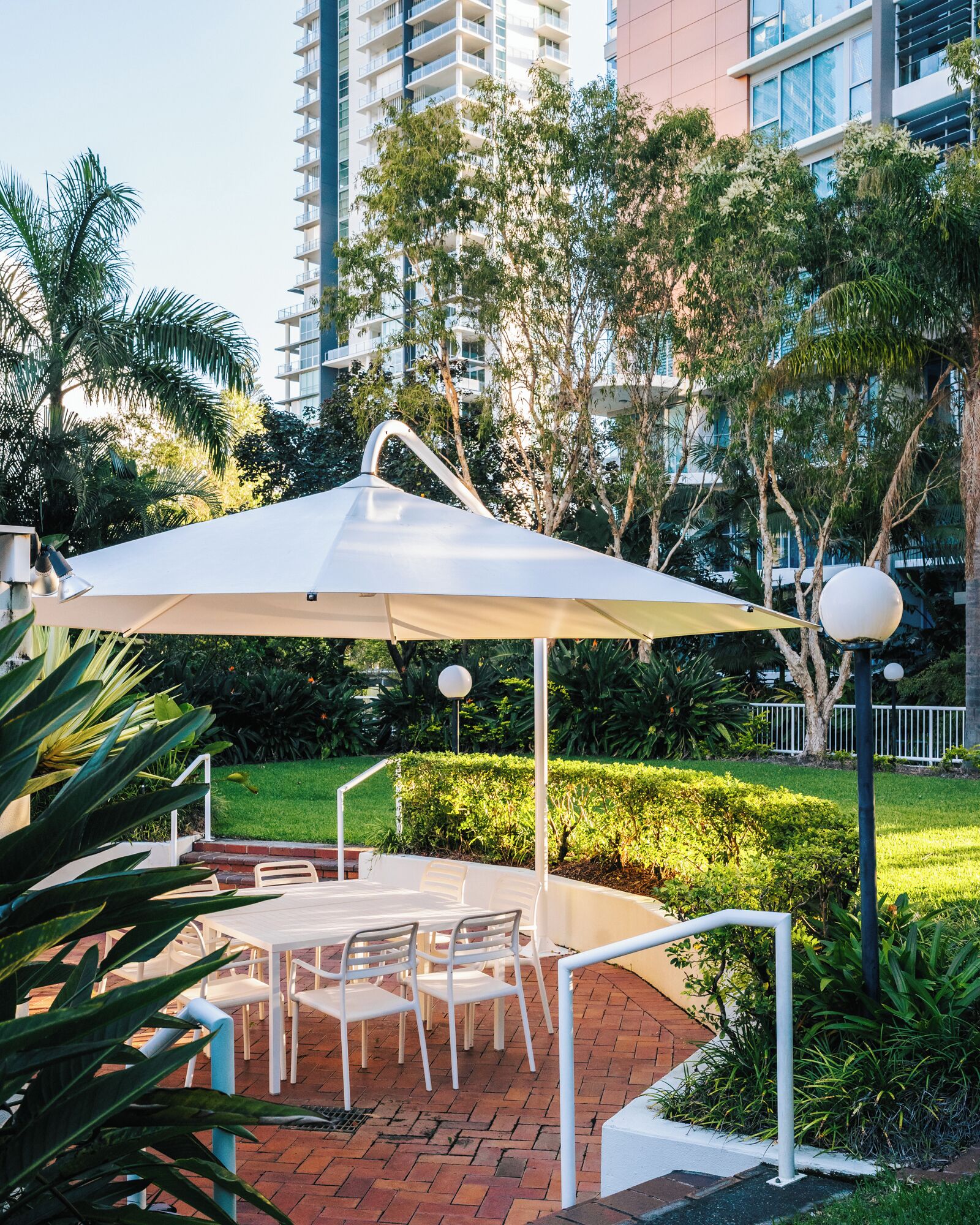 An outdoor patio with a table and chairs under a white umbrella, surrounded by greenery and a tall building in the background.