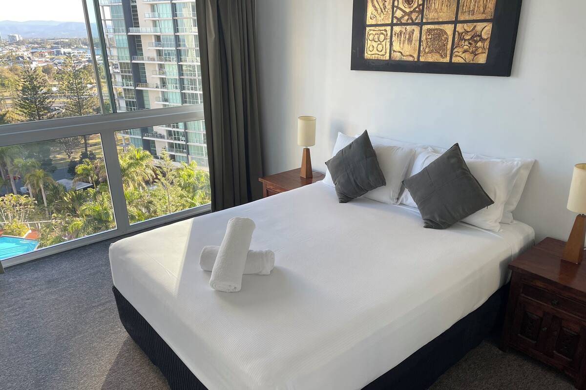 A hotel room with a large bed, two pillows, a neatly made bed, a view of a cityscape, a painting on the wall, and a lamp on the nightstand.