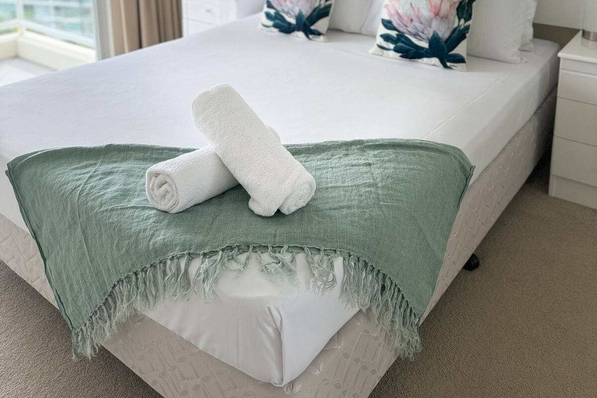 A neatly made bed with a green throw blanket and a rolled white towel, a white nightstand with a pink vase, and a painting of a landscape on the wall.