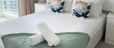 A neatly made bed with a green throw blanket and a rolled white towel, a white nightstand with a pink vase, and a painting of a landscape on the wall.