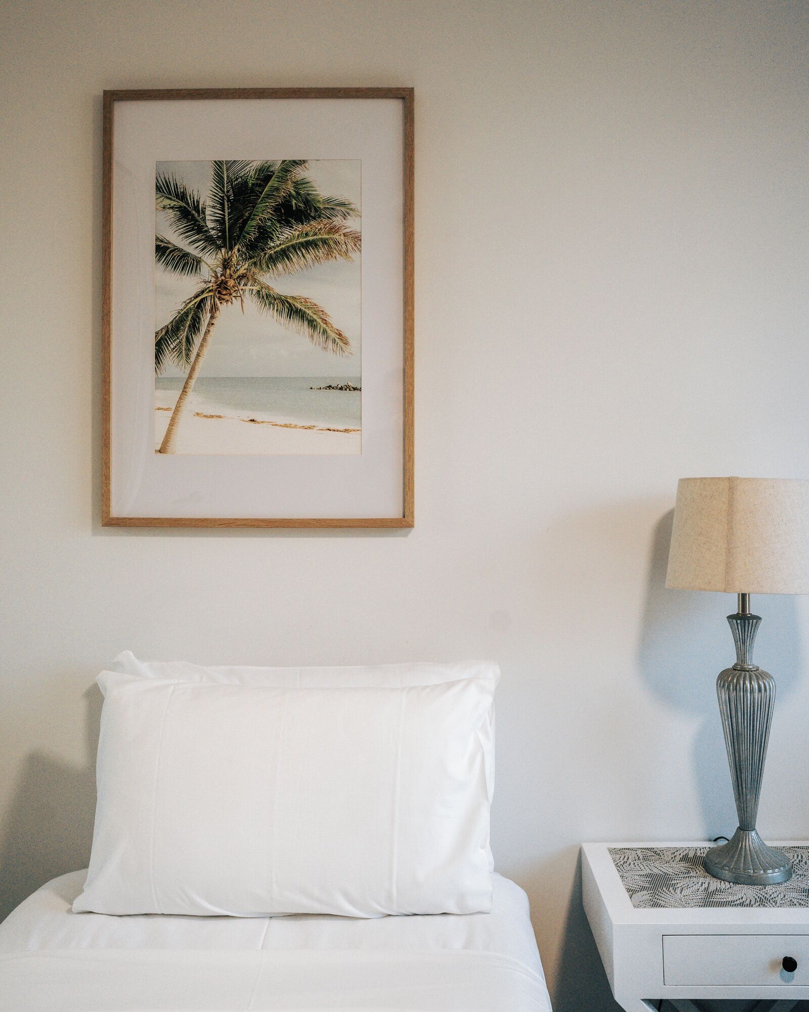 Un lit bien fait, avec du linge de lit blanc, un tableau encadré représentant un palmier accroché au mur et une lampe sur une table de chevet.