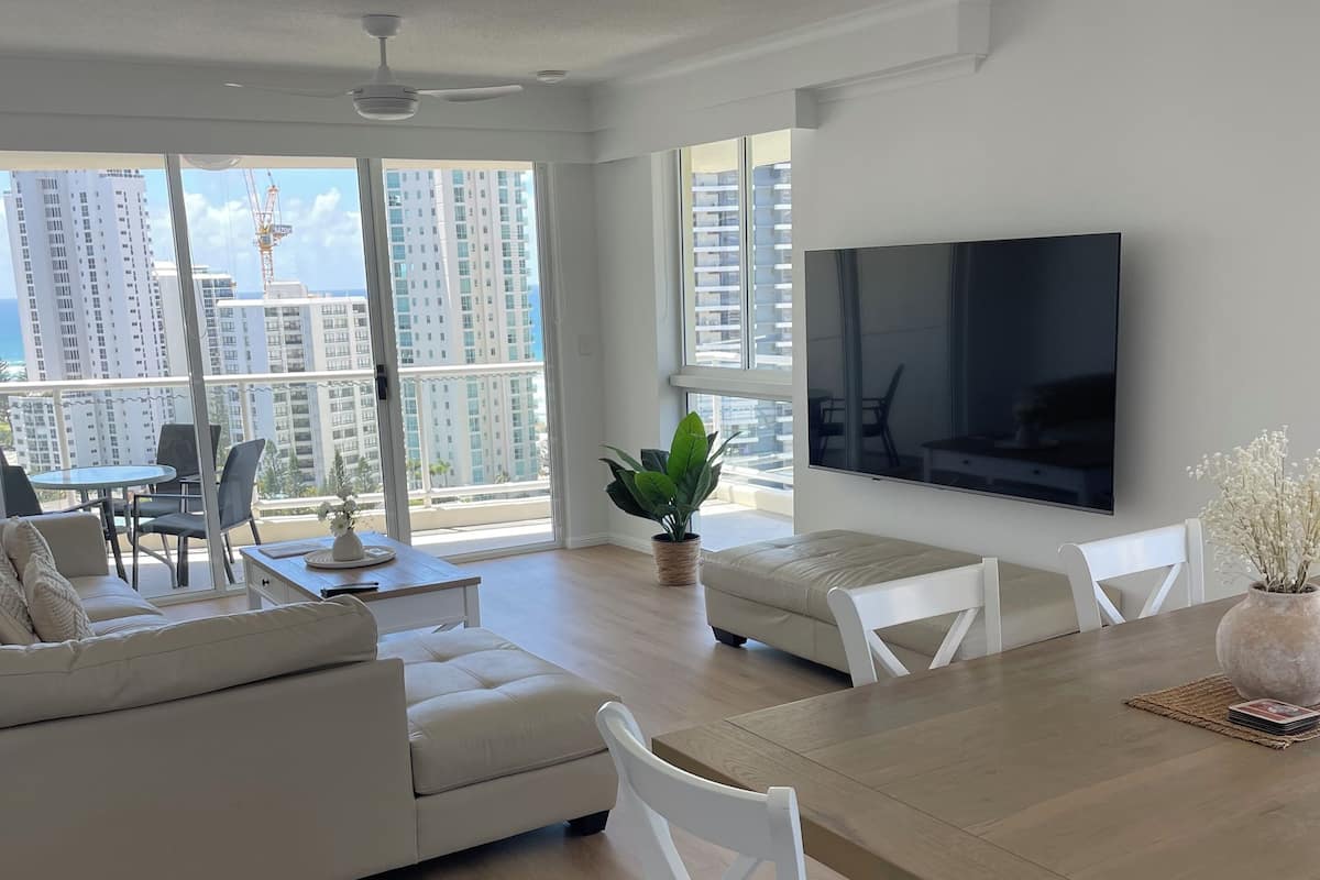 A modern living room with a large TV, white furniture, and a balcony with a view of high-rise buildings.