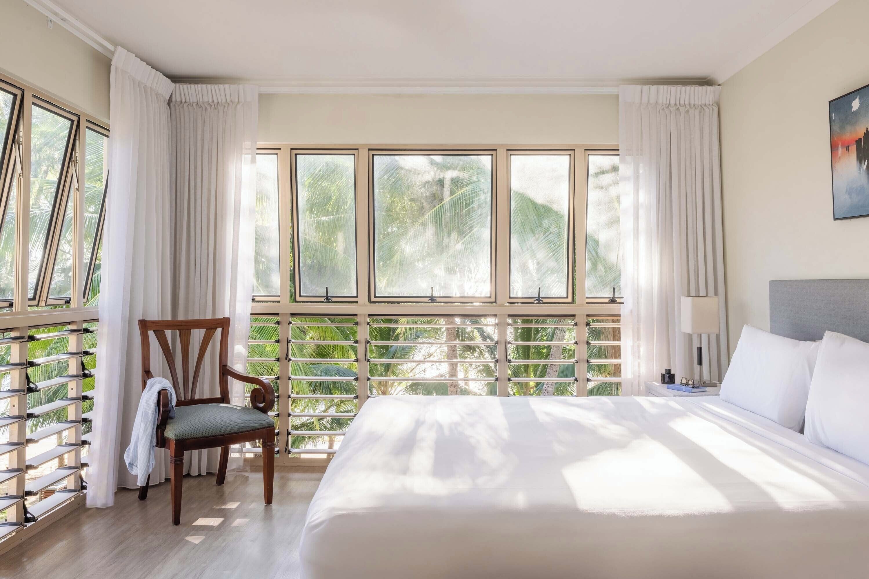 Hotellihuone, jossa on suuri sänky, tuoli, yöpöytä ja ikkuna, josta avautuu näkymä palmuille.