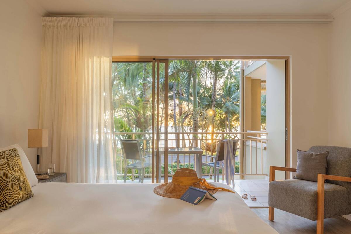 A hotel room with a bed, a chair, a nightstand, a lamp, a hat, and a book.