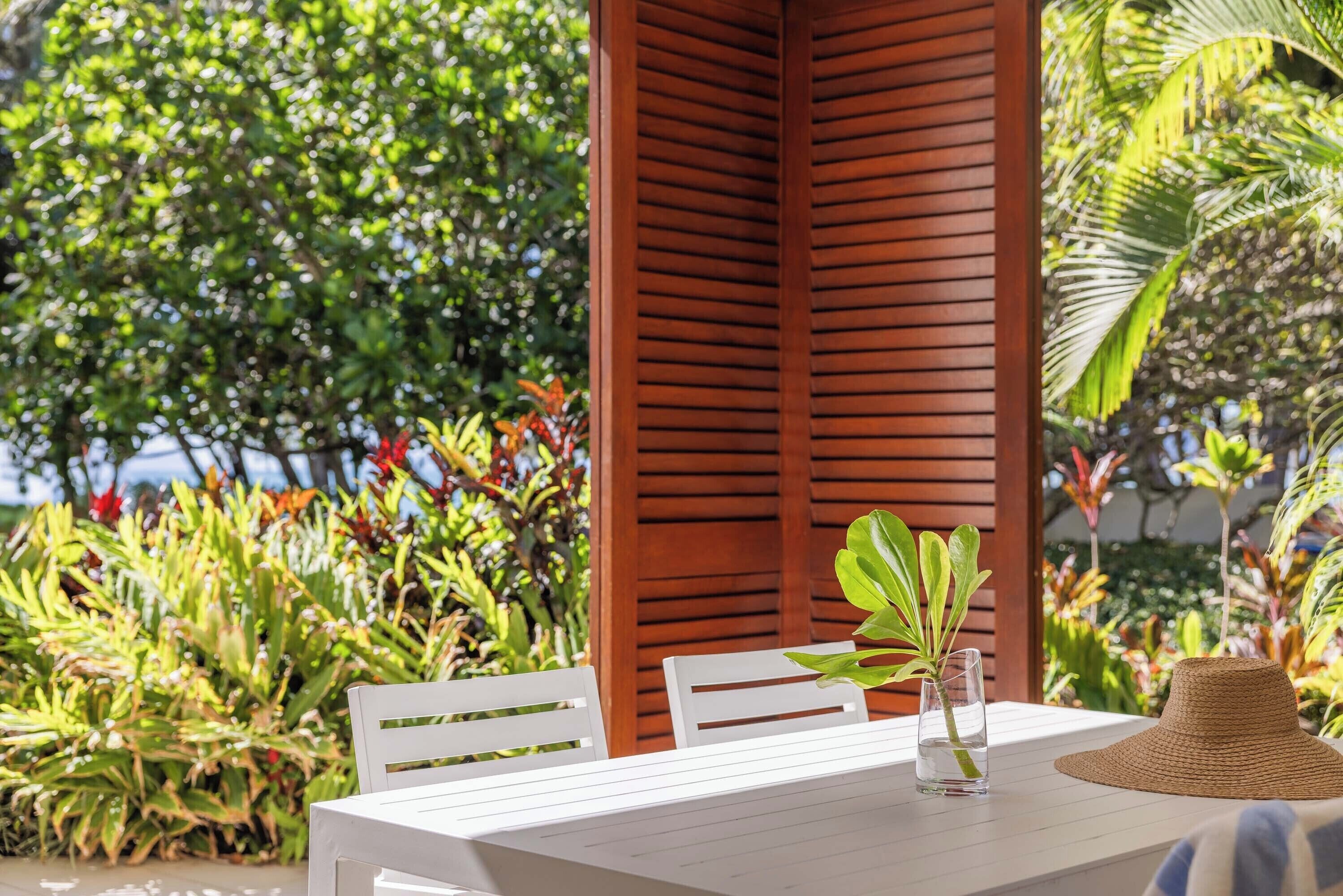 A white table with a glass of water and a straw, surrounded by white chairs, set against a wooden backdrop with lush greenery in the background.