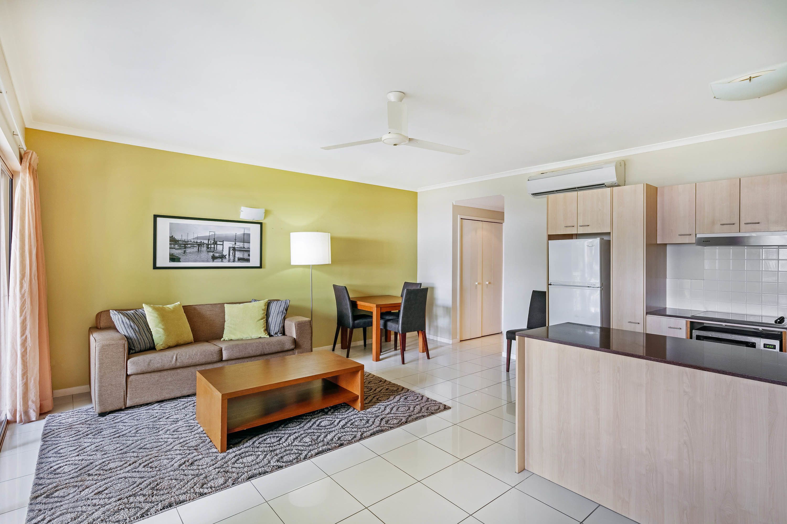 A modern living room with a sofa, coffee table, and dining area. There is a kitchen with cabinets and appliances.