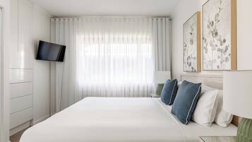 A modern hotel room with a large bed, a flat-screen TV mounted on the wall, and a large window with sheer curtains.