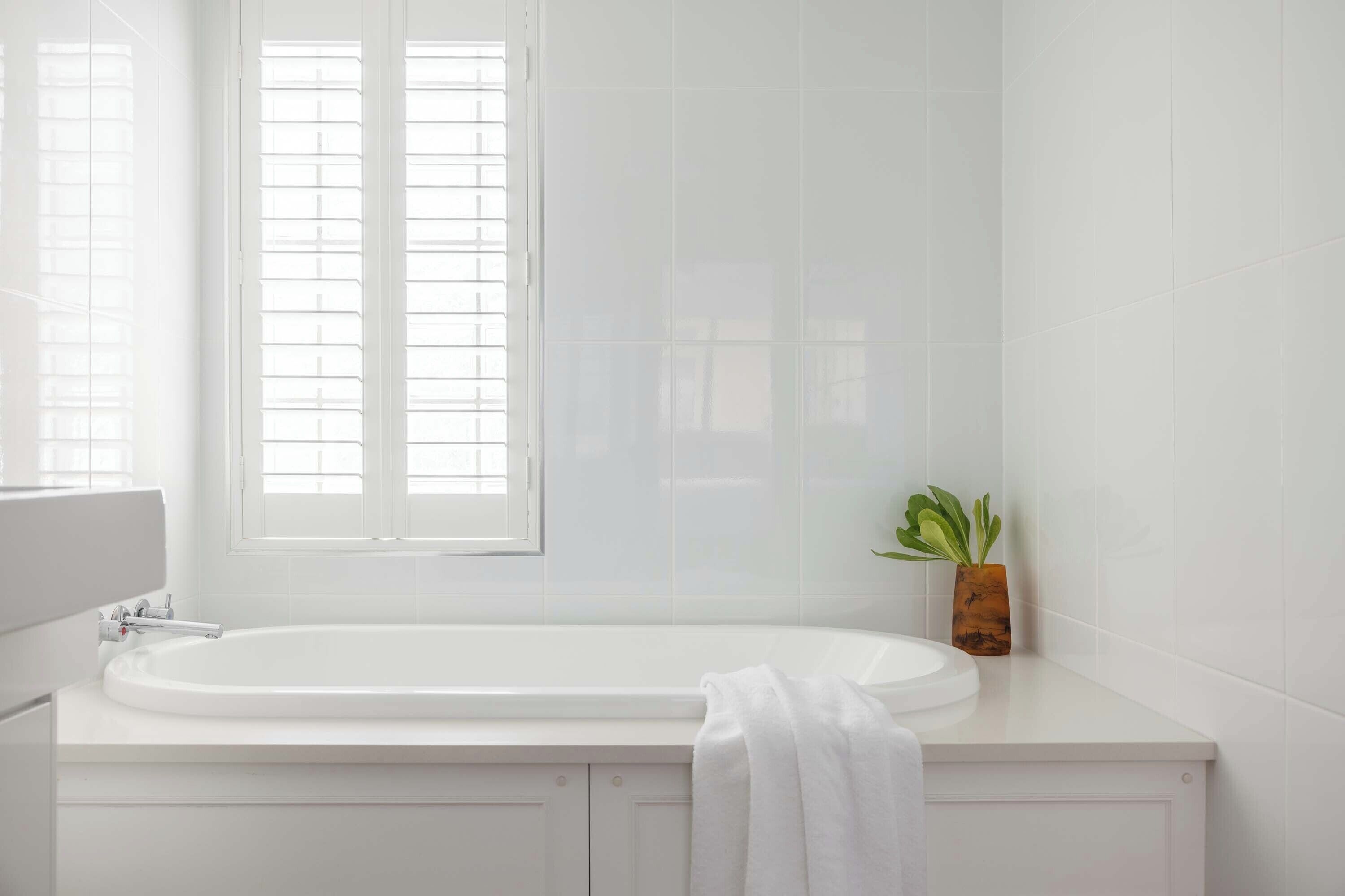 A modern bathroom with a white bathtub, a window with blinds, and a small plant on the counter.