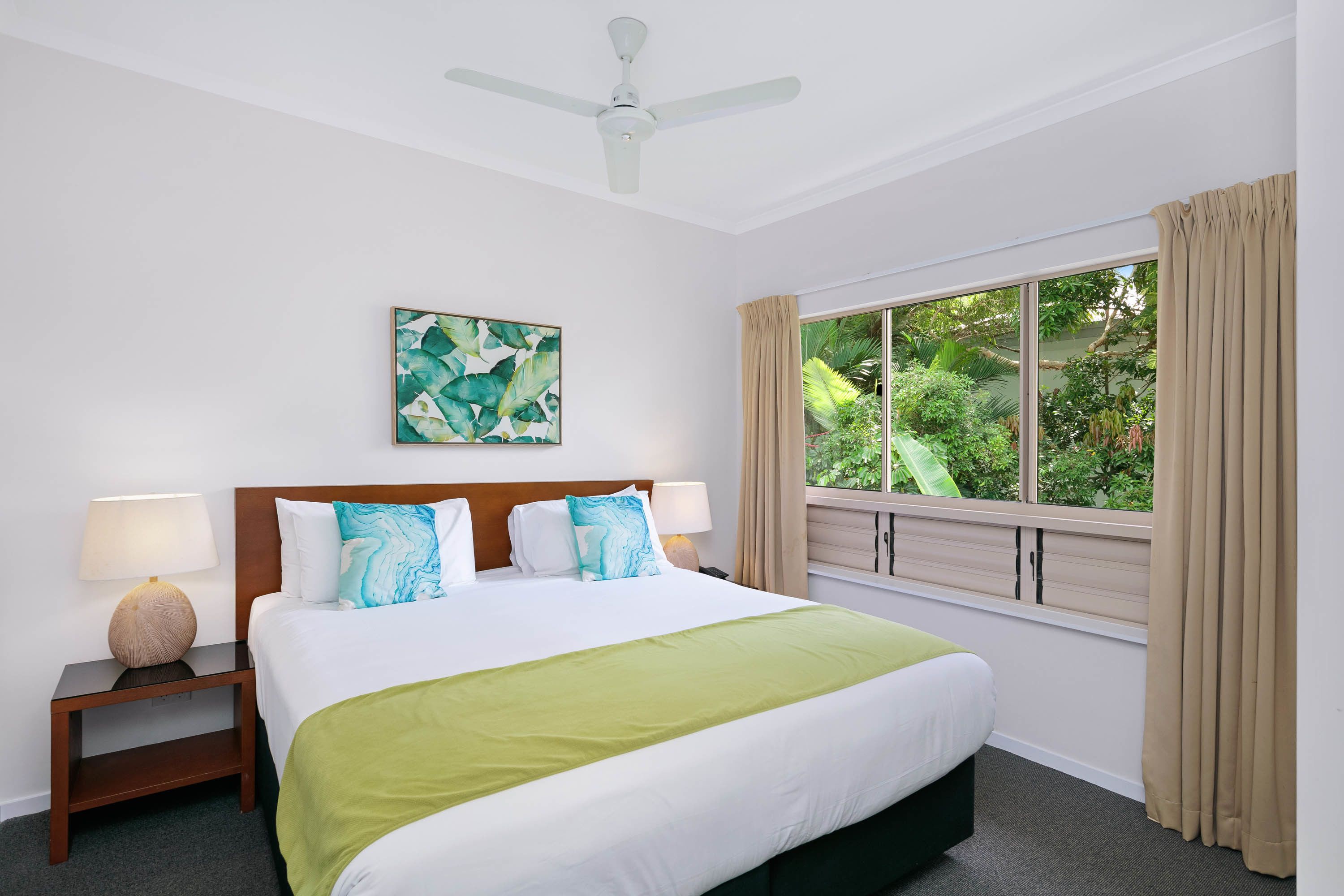 Une chambre d’hôtel avec un lit, deux tables de chevet, un ventilateur de plafond, une fenêtre avec des rideaux et une vue sur la verdure.