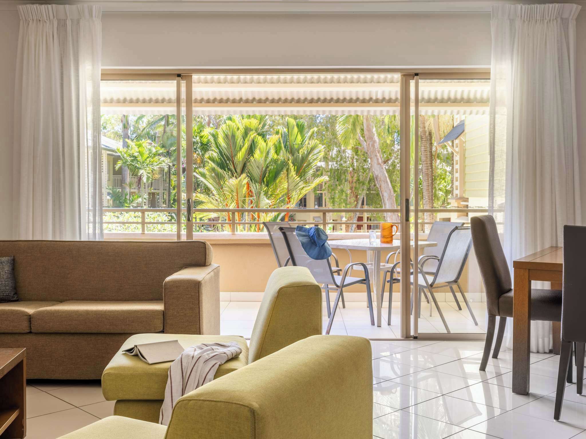 A modern living room with a sofa, armchairs, and a dining area with a table and chairs. Large windows offer a view of palm trees and a balcony.