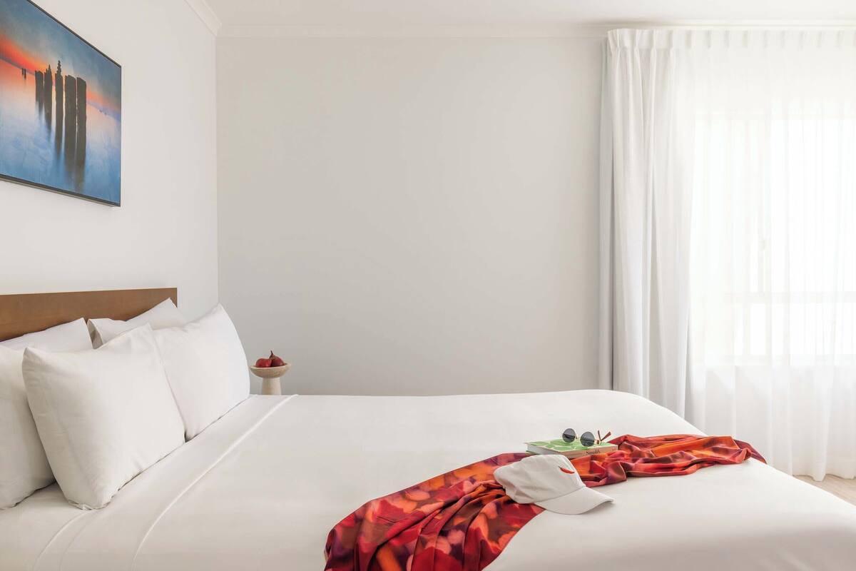 A neatly made bed with white bedding, a red and orange blanket, and a white pillow.