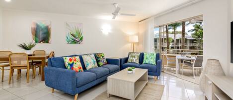 A modern living room with a blue sofa set, a wooden coffee table, and a dining area with wicker chairs.