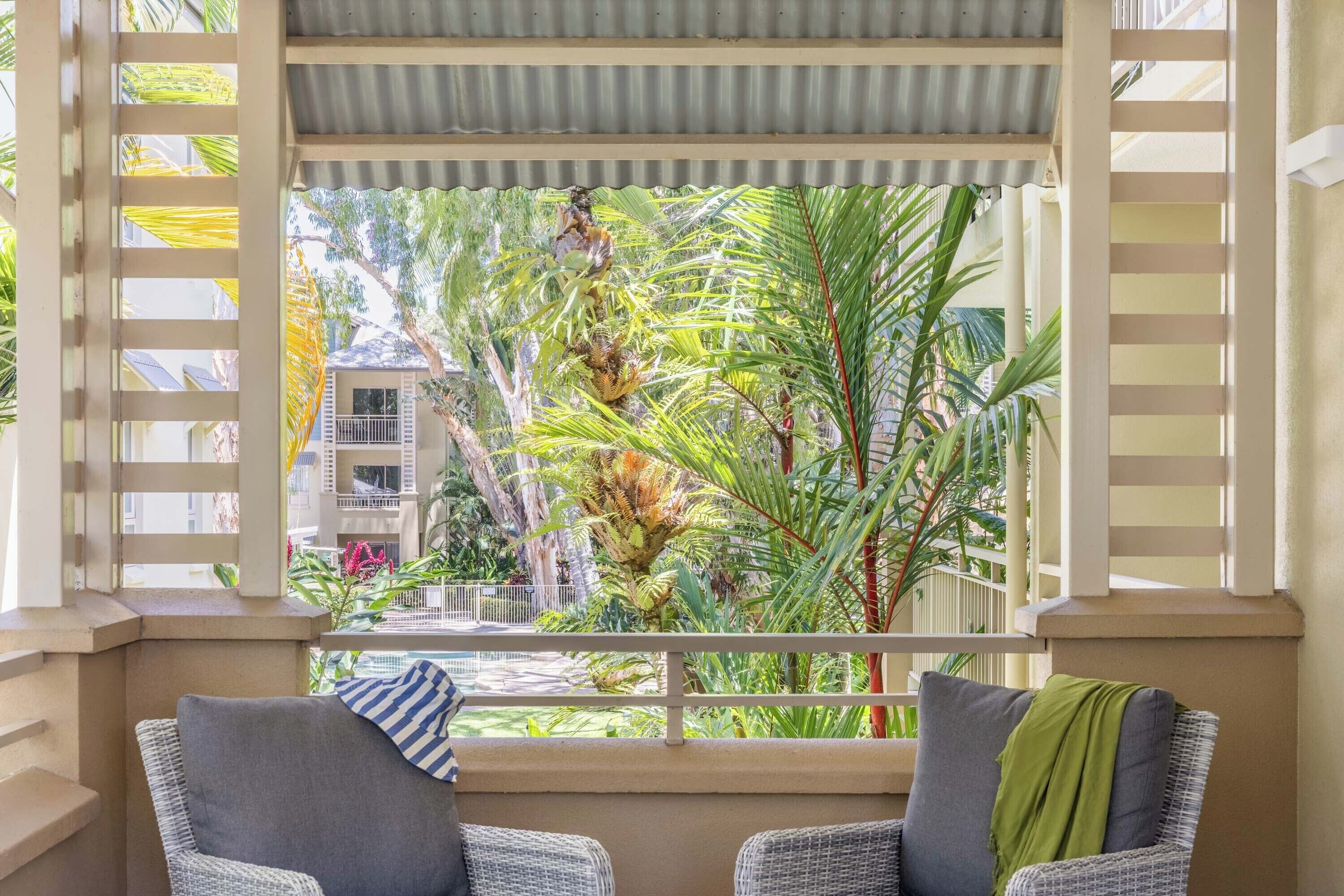 Een balkon met rieten stoelen, uitzicht op een tropische tuin en een gebouw op de achtergrond.