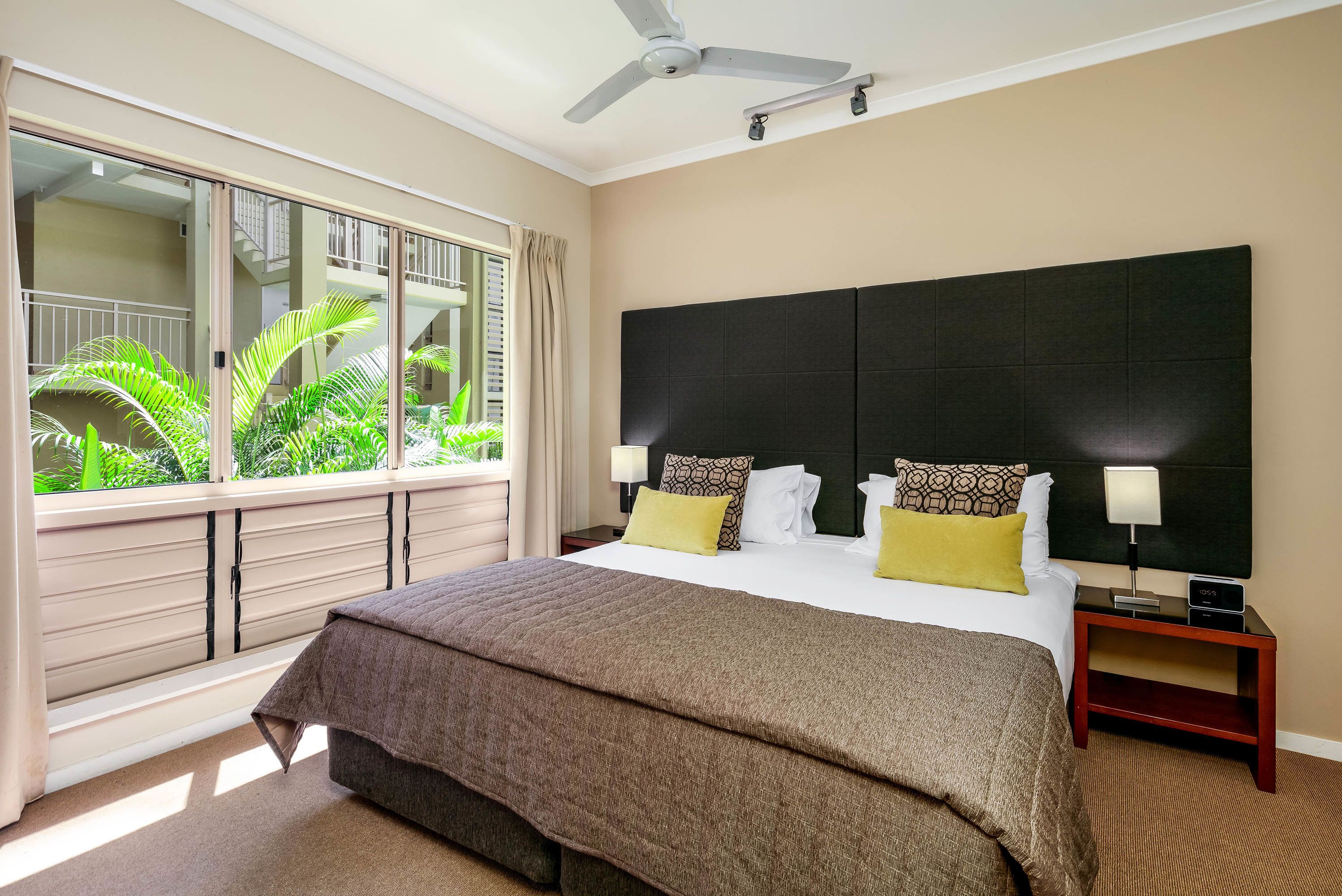 Una habitación de hotel con una cama grande, un ventilador de techo, una ventana con vista a zonas verdes y una mesita de noche con una lámpara.