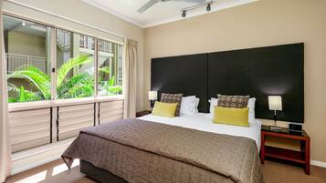 Una habitación de hotel con una cama grande, un ventilador de techo, una ventana con vista a zonas verdes y una mesita de noche con una lámpara.