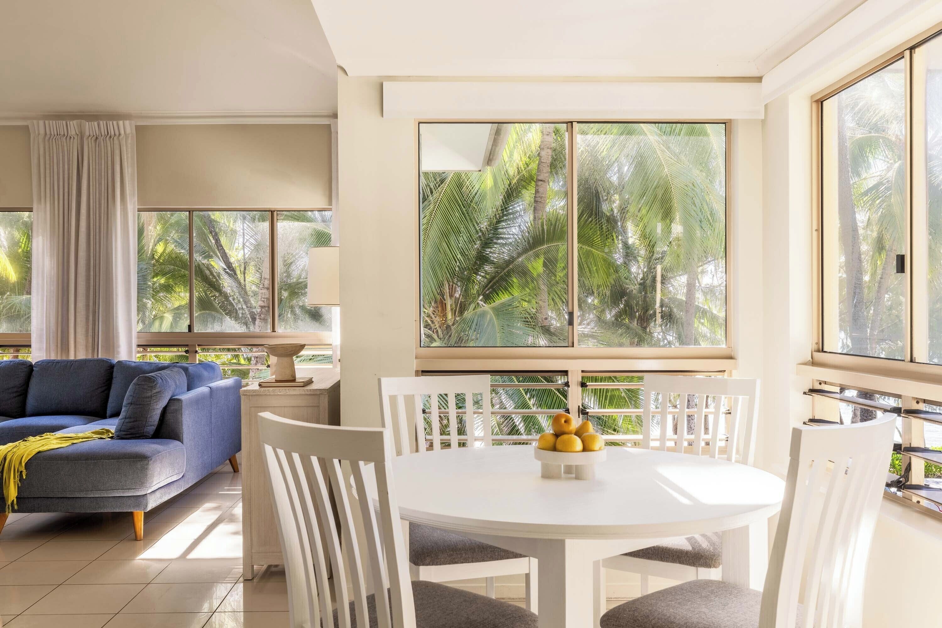 A modern living room with a blue sofa, white dining table, and chairs, large windows with a view of palm trees.