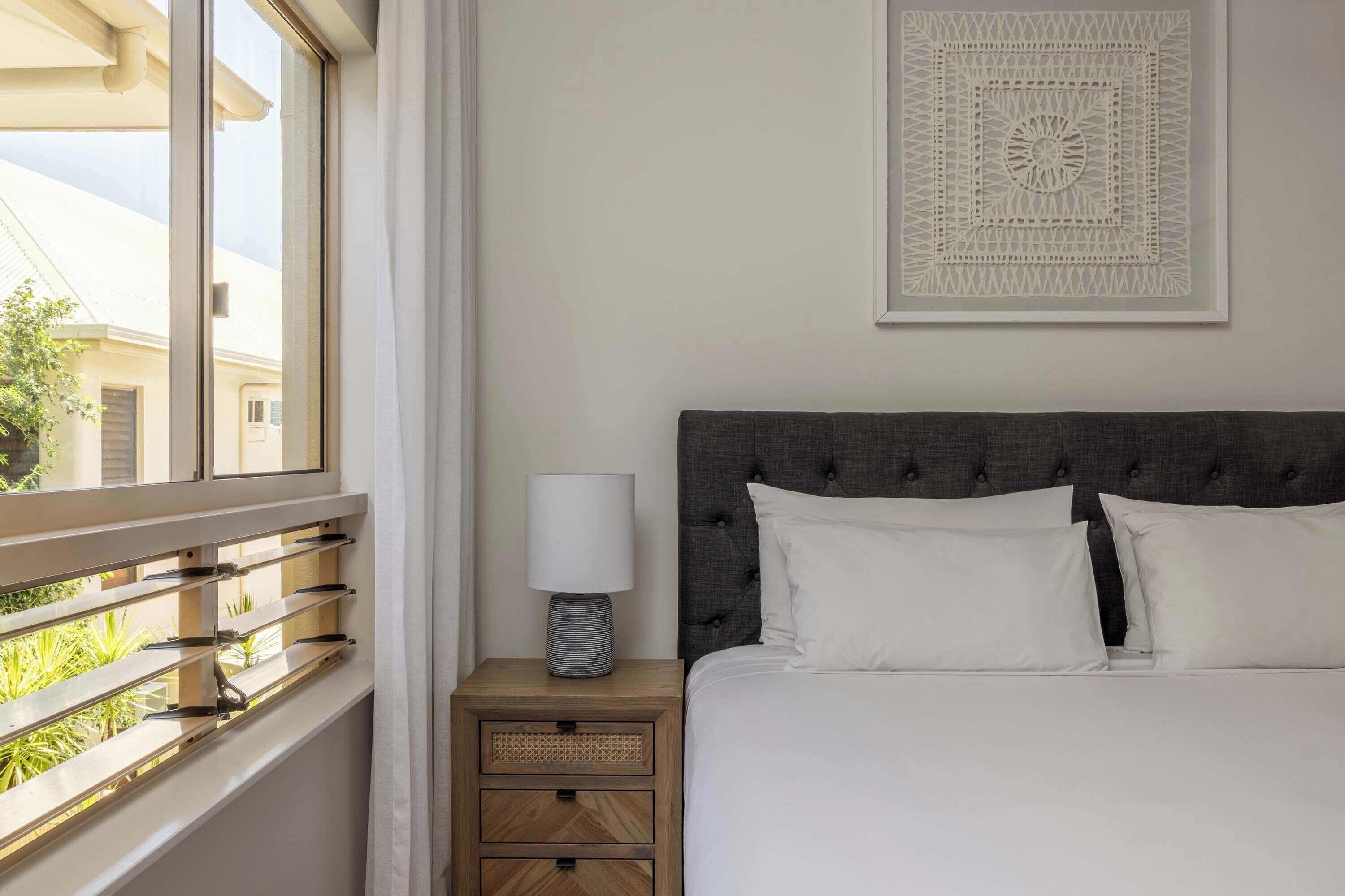 Una cama bien tendida con sábanas blancas, un cabecero tapizado en gris y una mesita de noche con una lámpara y un objeto decorativo.