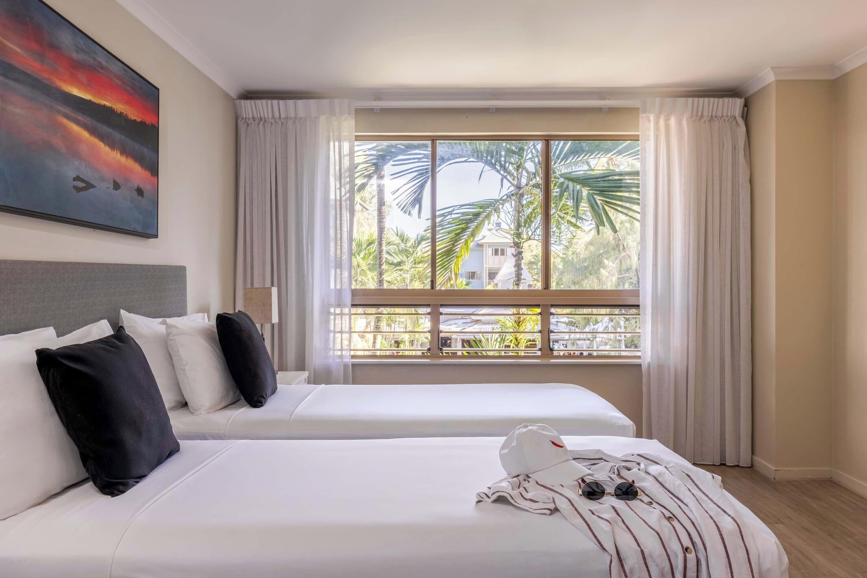 A hotel room with two beds, a large window with curtains, a painting on the wall, and a view of palm trees and buildings outside.