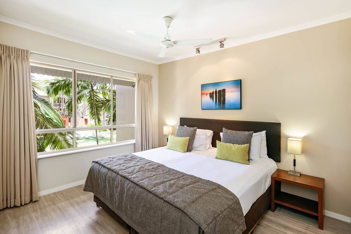 A hotel room with a large bed, a nightstand, a ceiling fan, a window with curtains, and a painting on the wall.