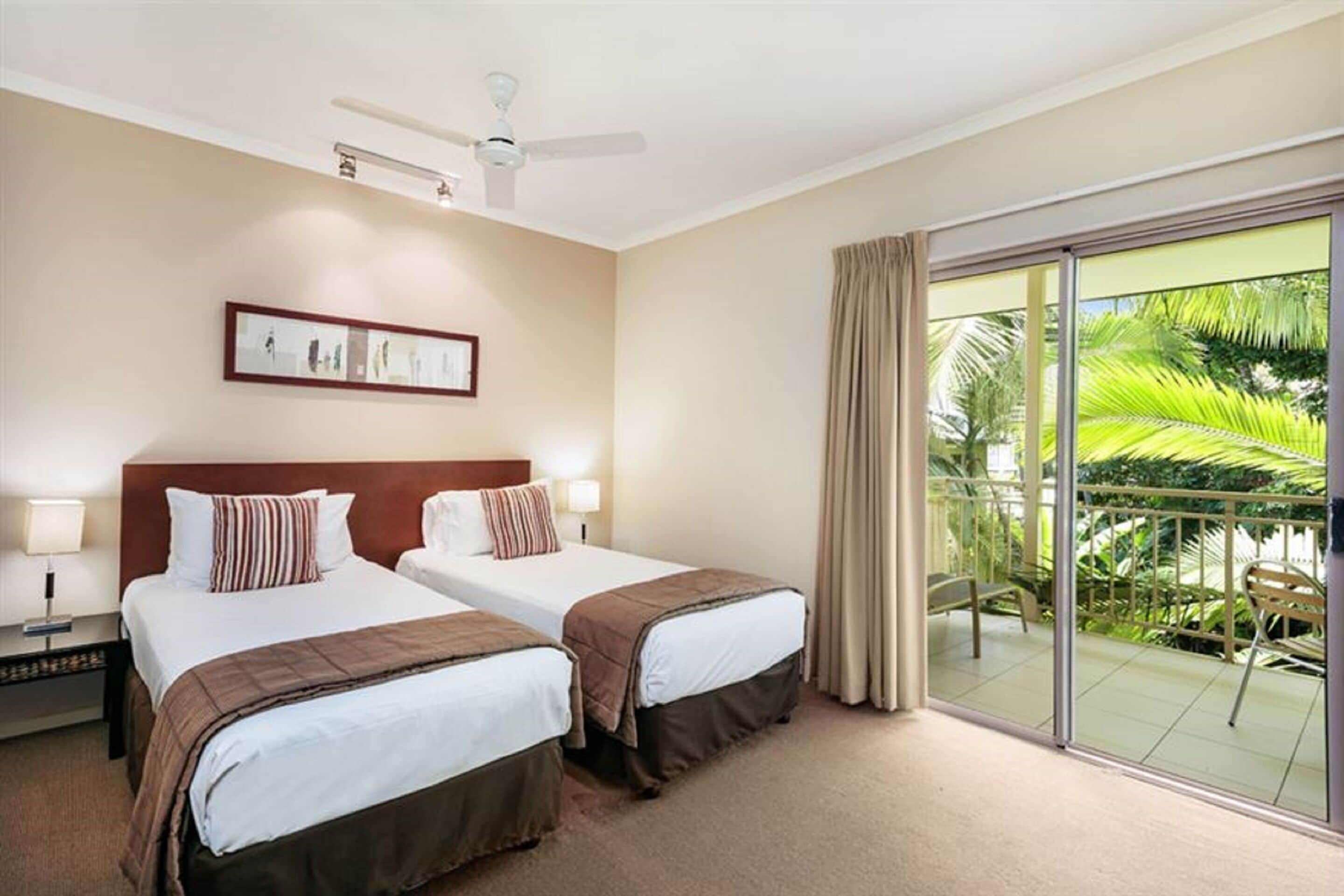A hotel room with two beds, a ceiling fan, a large window with a view of palm trees, and a framed picture on the wall.
