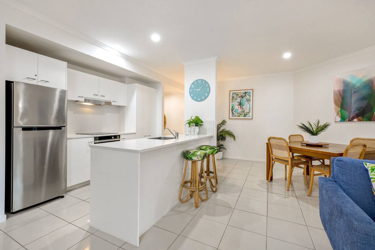 A modern kitchen with a built-in island, stainless steel refrigerator, and white cabinetry. There is a dining area with a wooden table and chairs, and a blue sofa.