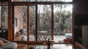 Uma cabana moderna com uma porta de correr de vidro que dá para um deck externo. No interior, há uma mesa de madeira com cadeiras, um churrasqueira e uma taça de vinho.
