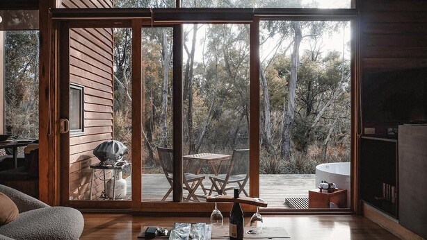 Una moderna baita con una porta scorrevole in vetro che dà su un patio esterno. All'interno, si trova un tavolo di legno con sedie, un barbecue e un bicchiere da vino.
