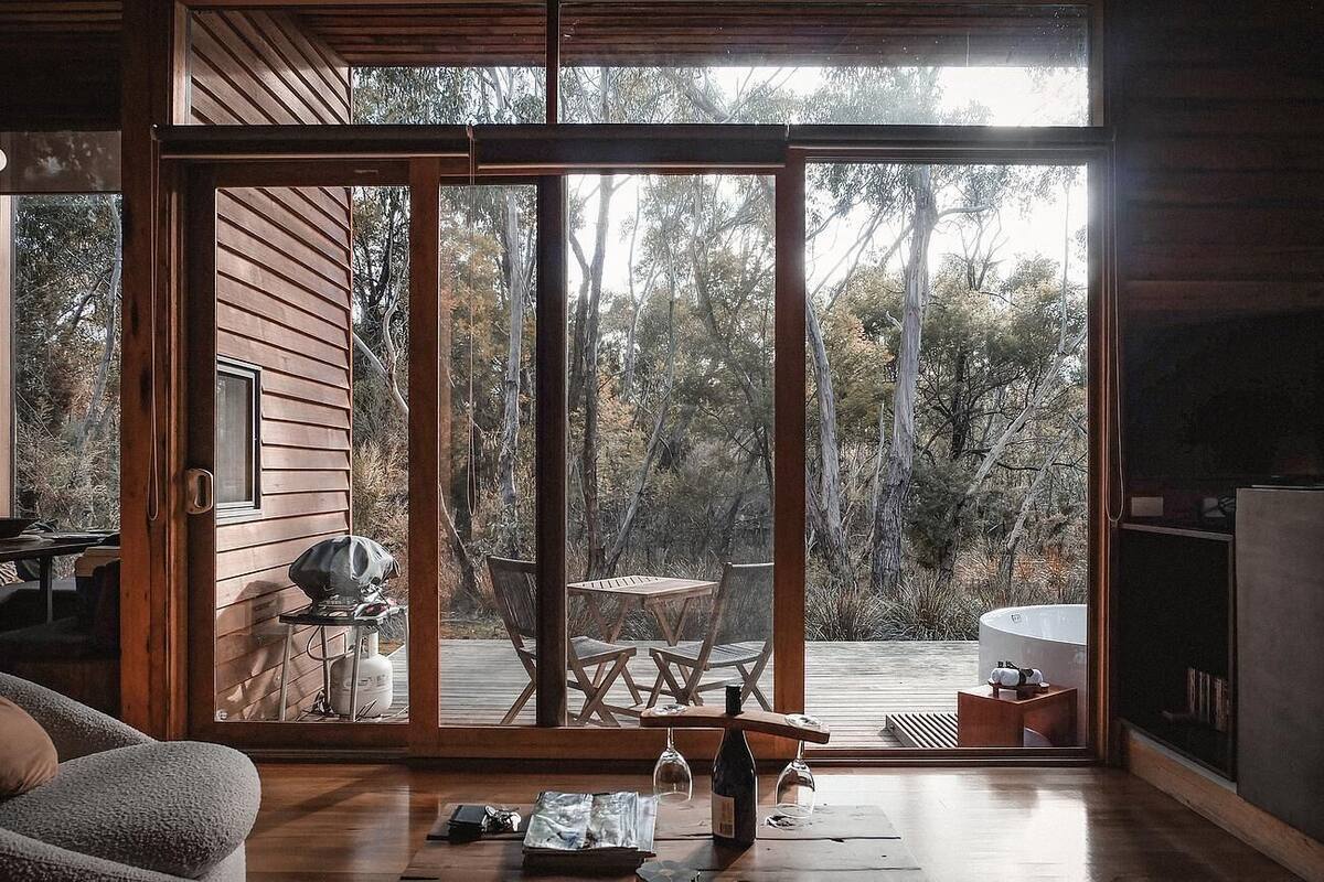 Una cabaña moderna con una puerta corrediza de vidrio que da a una terraza exterior. En el interior, hay una mesa de madera con sillas, una parrilla y una copa de vino.