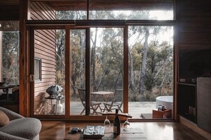Una cabaña moderna con una puerta corrediza de vidrio que da a una terraza exterior. En el interior, hay una mesa de madera con sillas, una parrilla y una copa de vino.