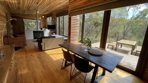 Un interno moderno di una baita con pavimento e soffitto in legno, un'area pranzo con tavolo e sedie, una cucina con televisore e ampie finestre che offrono vista sull'esterno.