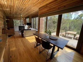 Interior moderno de cabaña con pisos y techo de madera, zona de comedor con mesa y sillas, cocina con televisor y ventanas amplias que ofrecen vistas al exterior.