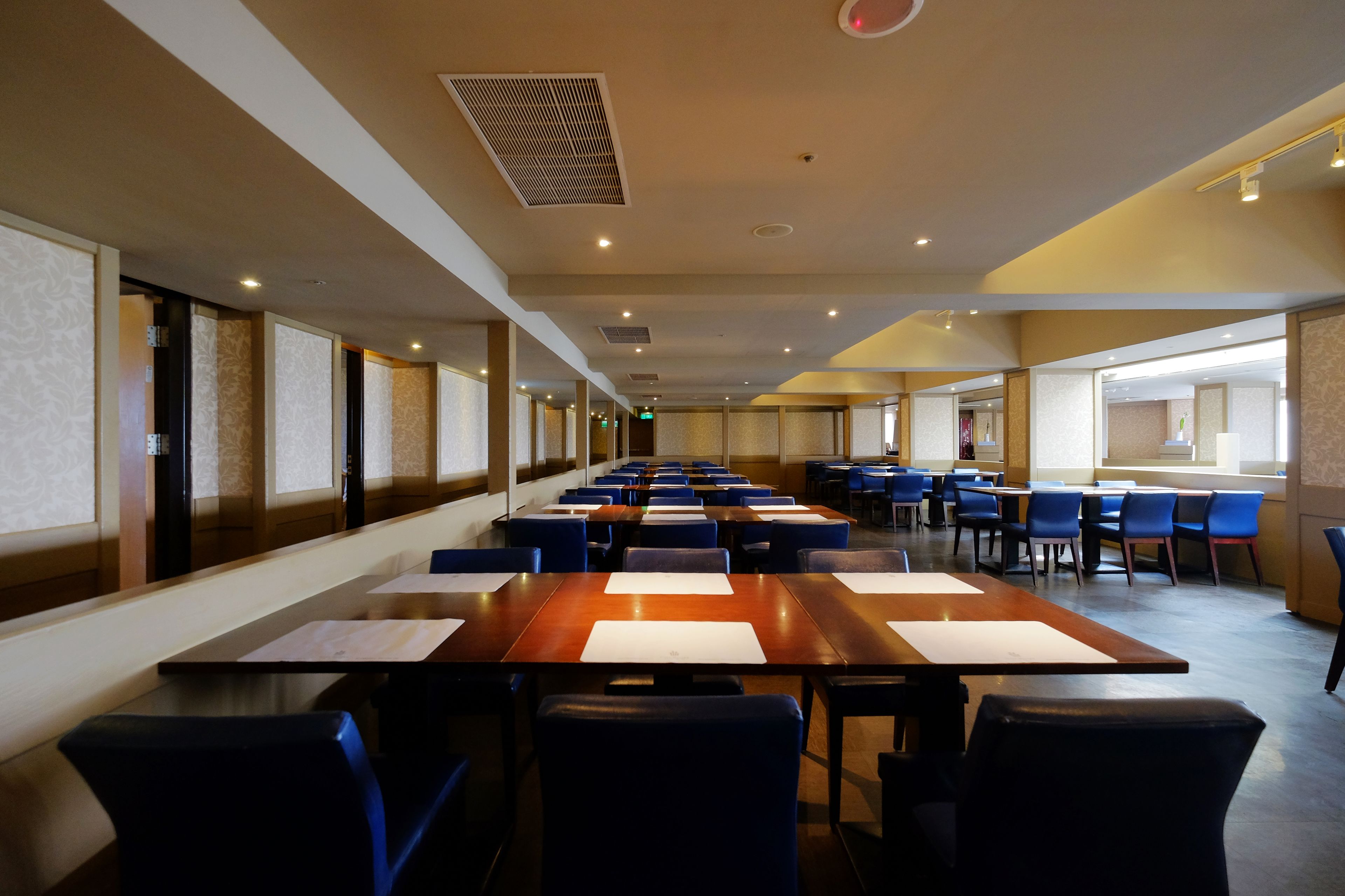 Un salón de conferencias con mesas y sillas largas, ventanas grandes y un techo alto con ventilación.
