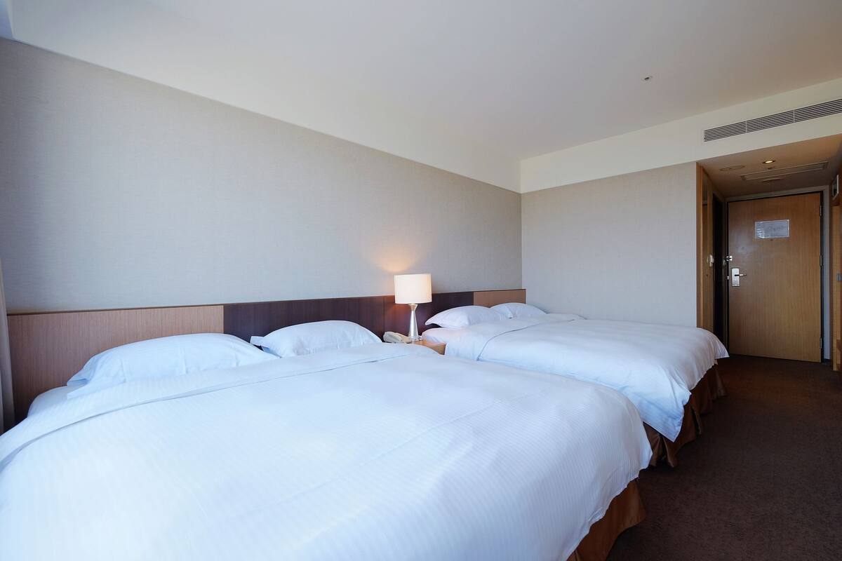 Een hotelkamer met twee bedden, een nachtkastje met een lamp, en een deur aan de rechterkant.