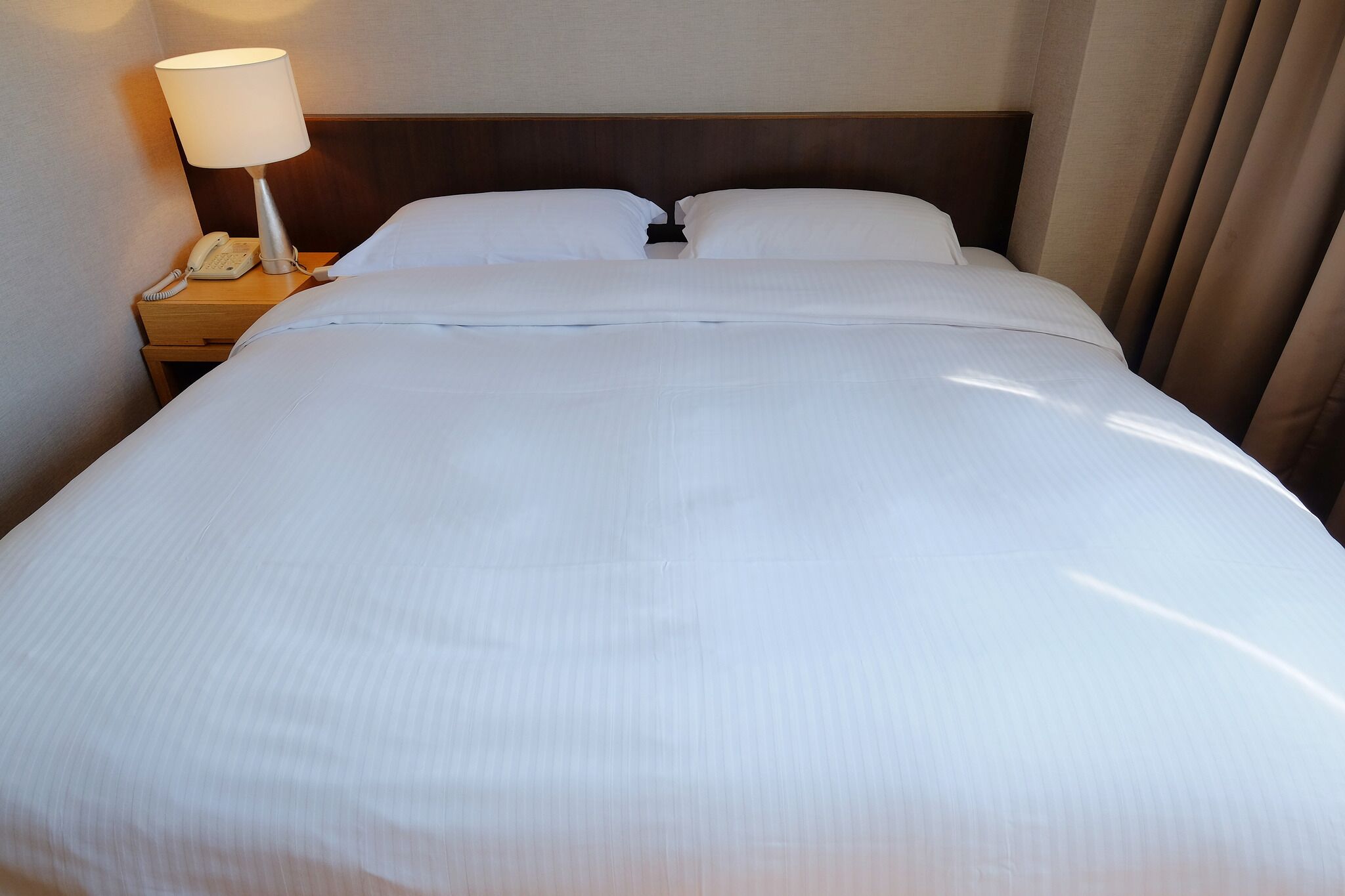 Una cama bien hecha con sábanas blancas, una lámpara de noche y un teléfono sobre una mesita de madera.